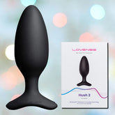 A black Lovense Hush 2 Vibrating Butt Plug - Medium is positioned beside its packaging box. The box features an image of the product along with branding details and specifications on a blurred, multicolored background, promising comfortable wear and intense vibrations.